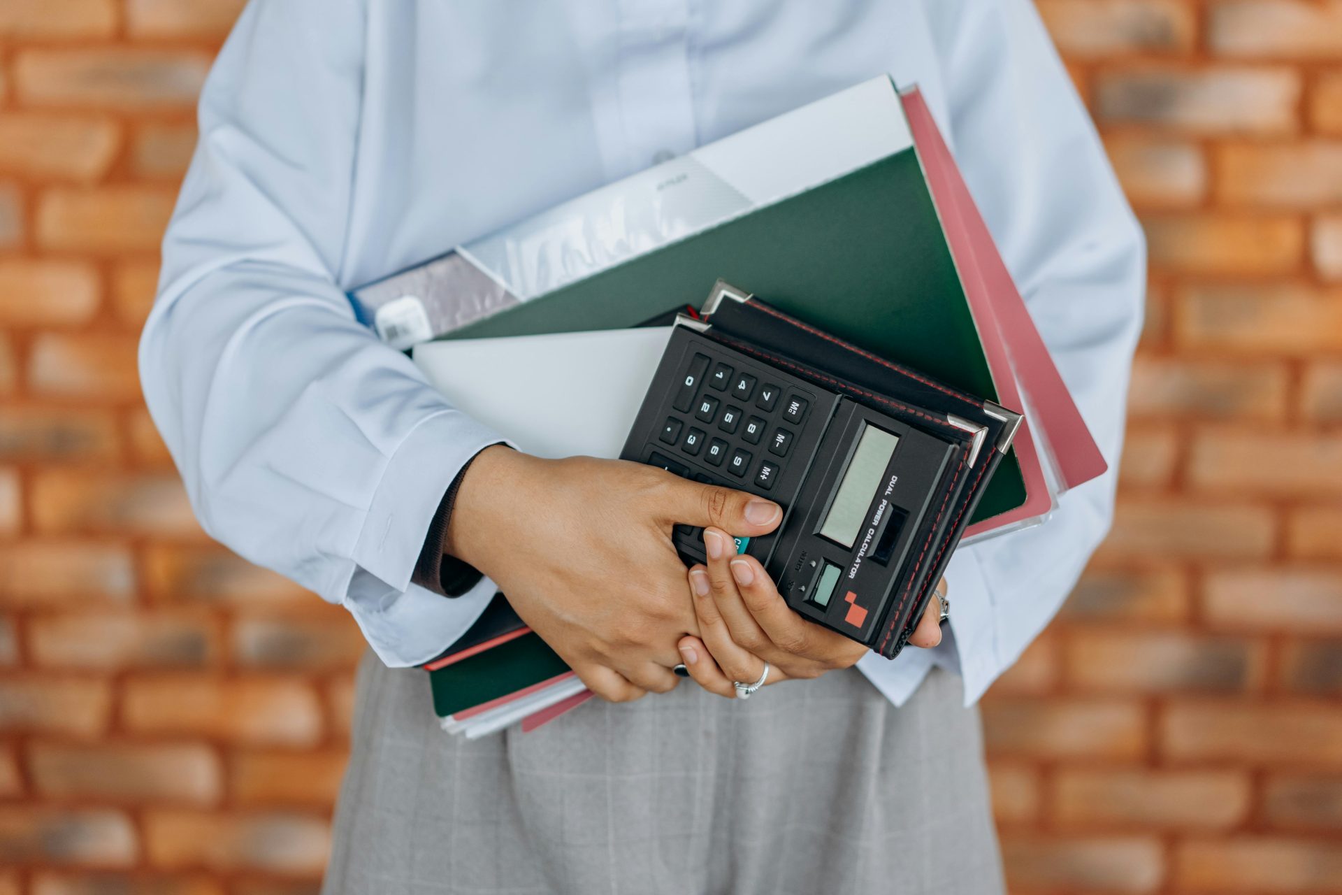 Gros plan d'une personne tenant une calculatrice et des dossiers sur fond de mur de briques.